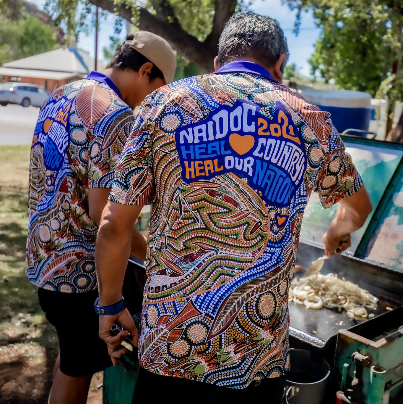 Two men wearing NAIDOC shirts cooking on a barbecue